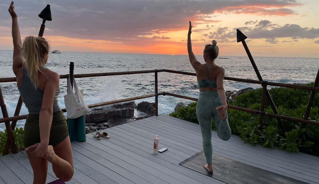 women doing yoga on beachfront deck overlooking ocean during sunset