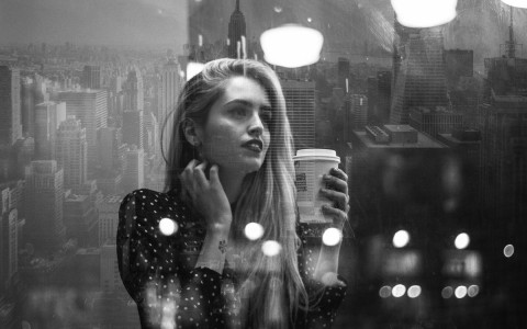 woman watching her reflection on a window while drinking coffee