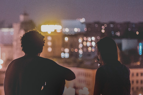 a man and woman sitting on a rooftop at night looking at buildings