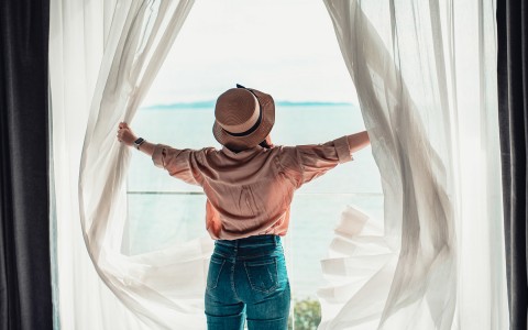 woman opening white curtains and looking out into the ocean