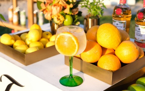 a glass filled with a fruity beverage and an orange slice in front of baskets of oranges and limes