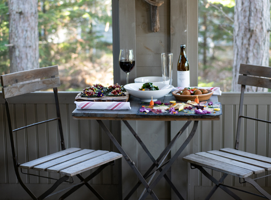 two chairs and a table on a porch with dinner set up and some red wine