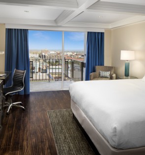 King room with crisp white sheets, shared night stand and lamp. Lounge chair and large window is adjacent to bed. Desk, dresser and mounted tv on opposing wall from beds