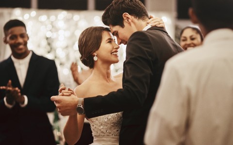 a smiling bride and groom dancing as guests watch and clap with soft lighting