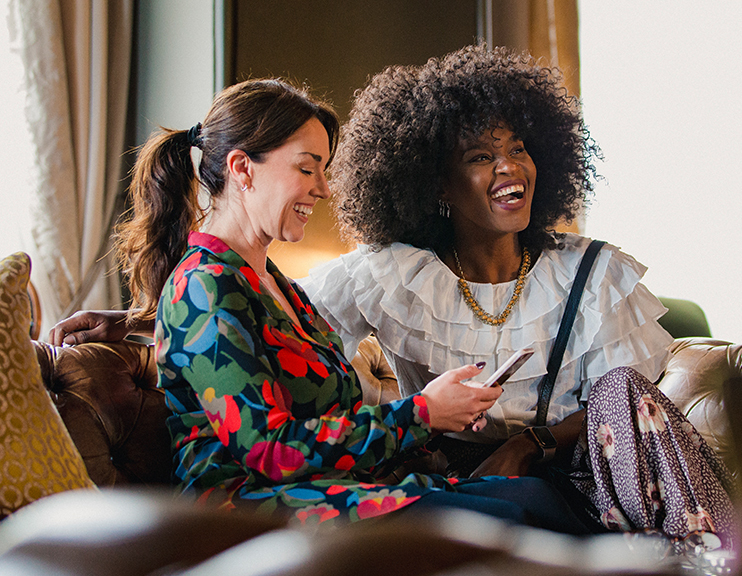 an african american woman and a white woman sitting by the sofa laughing