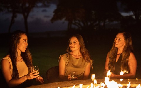 Three women charing and drinking next to a wood fire
