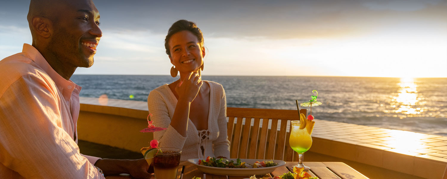 A woman looking at her boyfriend and drinking some tropical beverage at a romantic sunset