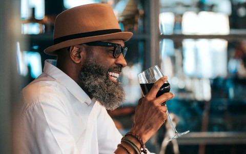 man in a hat wearing sunglasses drinking a glass of red wine at a bar smiling
