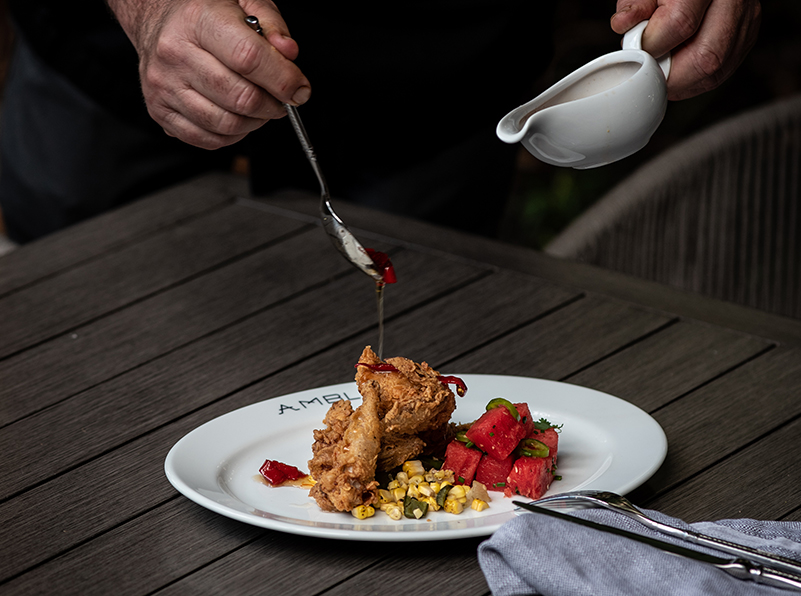 chef pouring sauce on fried chicken on a plate with watermelon and corn salad