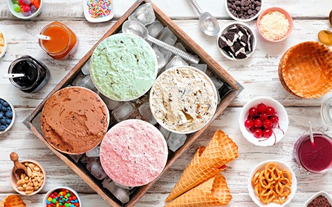 view of tubs of ice cream flavors with toppings and cones surrounding 