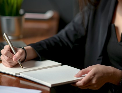 A woman wearing a black suit writing in a notebook at an oak desk with a laptop to the right.