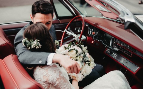Groom and bride sitting in an old fashioned convertible leaning in for a kiss