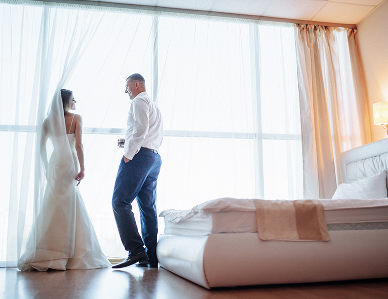 A smiling bride and groom in a room standing at the windows looking at each other.