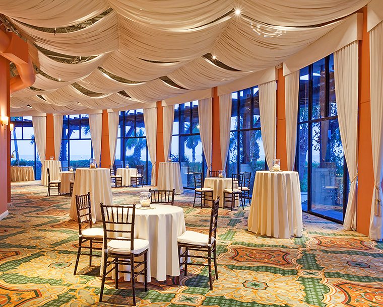 A mixture of cocktail tables and seating tables in an event room with an wall full of windows. White curtains are draped across the ceiling and gather in between each window.