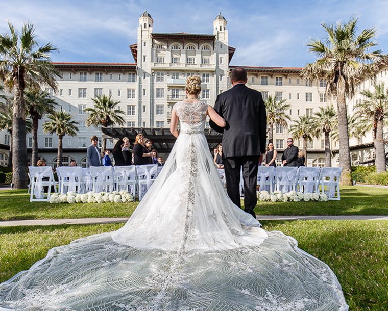 Behind view of a man walking his daughter down the isle at her wedding on the lawn outside Hotel Galvez