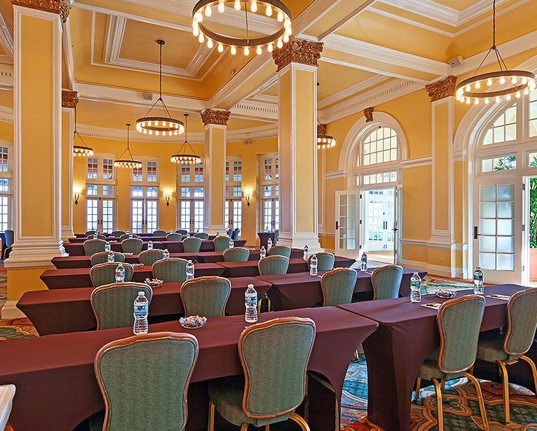 An large event space with yellow walls and white crown molding and rows of long tables draped in maroon tablecloths and blue cushioned chairs