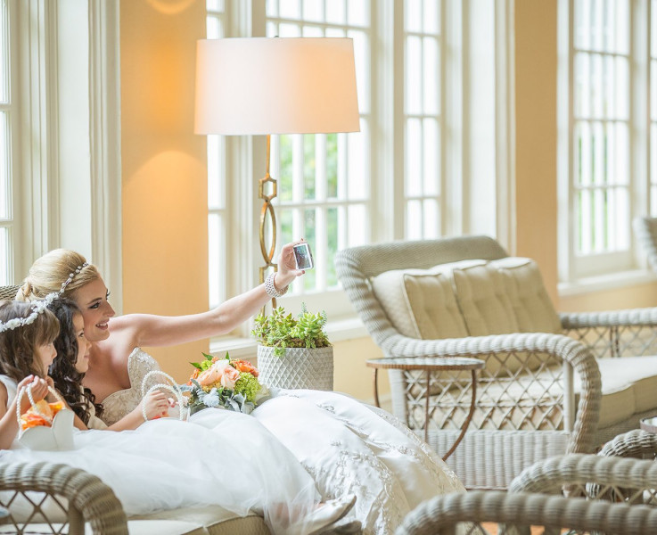 A bride sitting on a wicker couch taking a selfie with the flower girl 