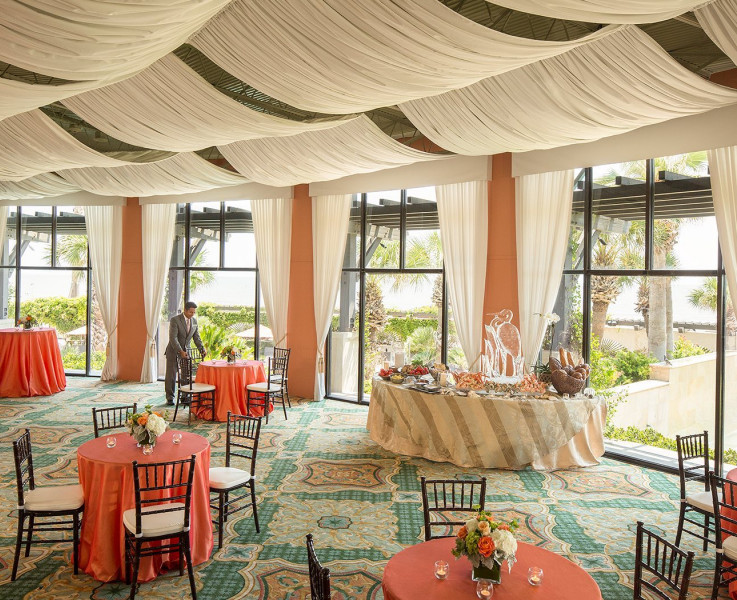 A colorful event space with cocktail tables, seating tables, and a buffet table. The ceiling is draped with white curtains that gather at the windows on the wall
