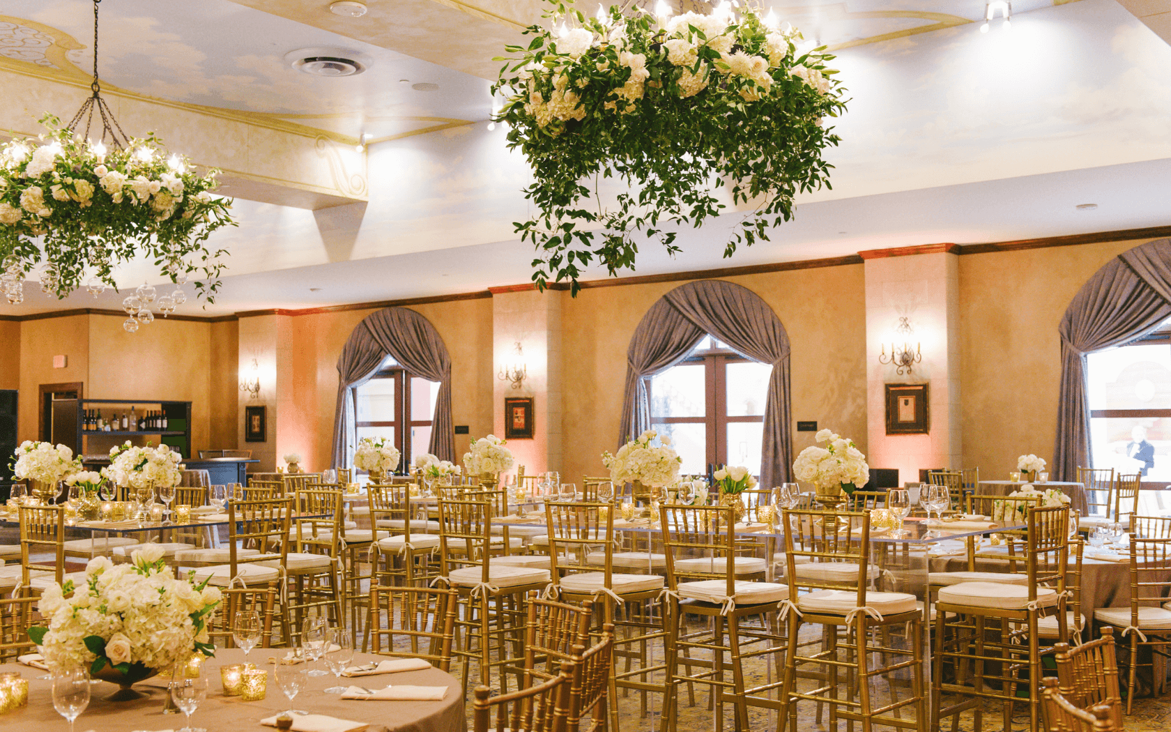 ballroom wedding reception with gold chiavari chairs, white floral arrangements, greenery hanging from chandeliers