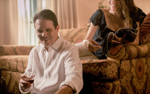 man with a glass of wine and his lady sitting on the sofa behind him with a magazine