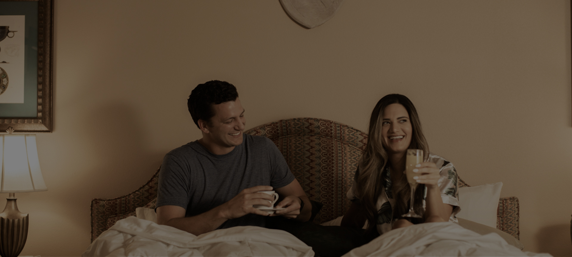 couple in bed smiling and laughing as they sip on their drinks