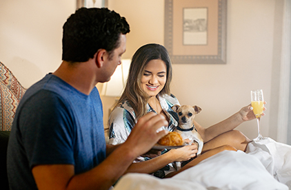 couple in bed with their small dog and a the lady holding a mimosa
