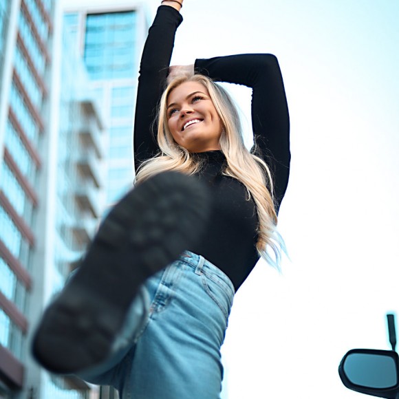 woman in black long sleeve, blue jeans, and black boots smiling and kicking toward the camera