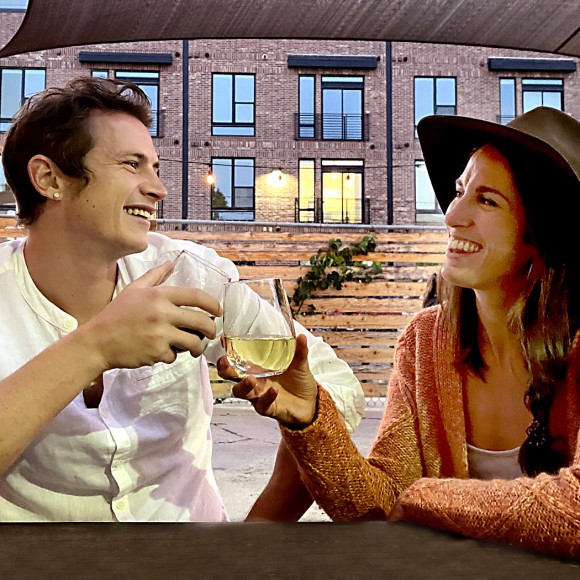 couple sitting on rooftop terrace smiling at one another and toasting with white wine