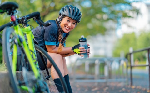 woman in biking gear with her bike drinking water on a bench outside