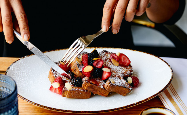 Person cutting into a french toast covered in strawberries, blueberries, almonds and powdered sugar