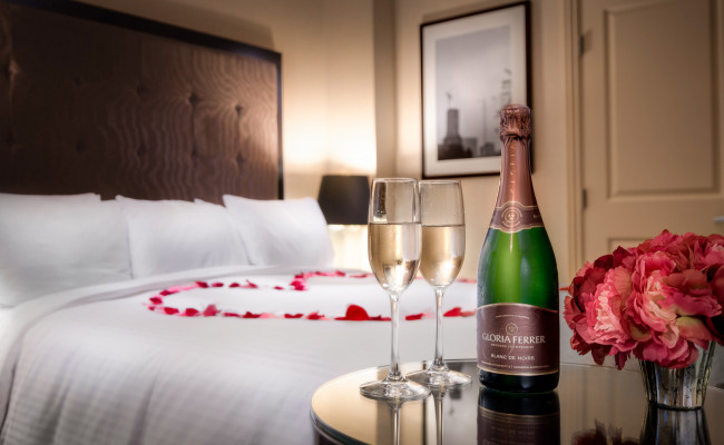 Bottle of champagne and two champagne glasses on a table with flowers, in front of a hotel bed with rose pedals in the shape of a heart