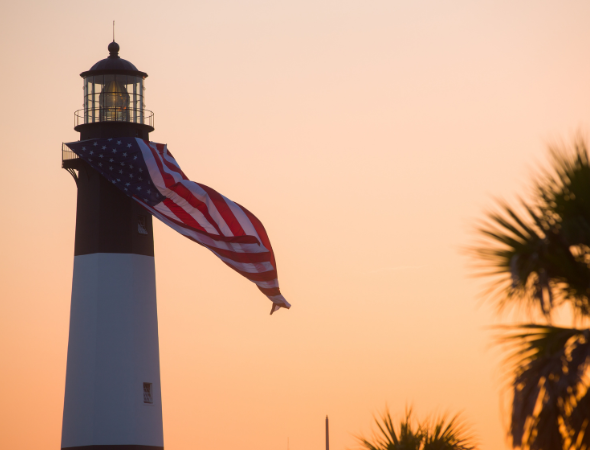 lighthouse with an american flag hanging off of it with a sunset in the background