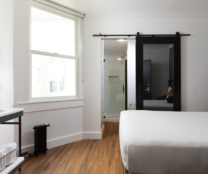 Corner view of a hotel room with features as queen bed, sink, small mirror, large mirror on the wall and a private bathroom