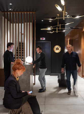 Concierge checking in a man while another man leaves the hotel with a suitcase and a red head woman sitting down texting