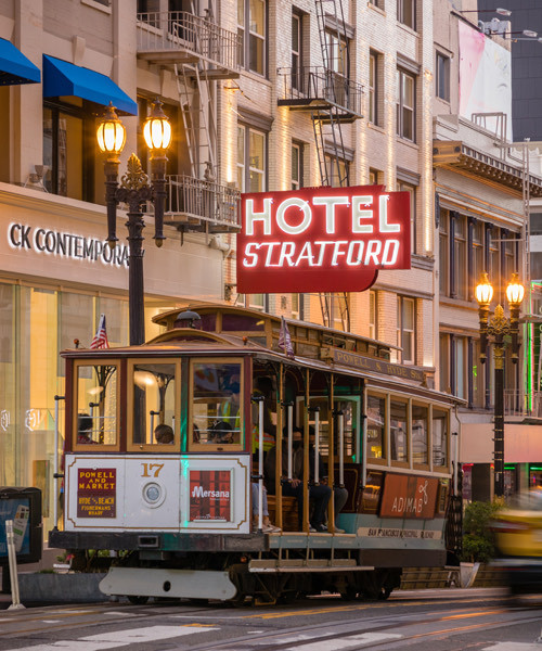View of hotel from outside with with a big red neon sign that says hotel stratford in white while trolly rides by