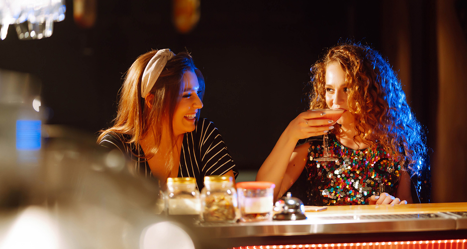 woman laughing looking at another woman while she is having a sip in a bar 