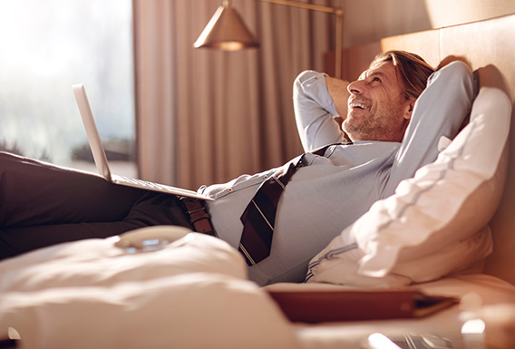 well dressed man laying on top of a guest bed with his laptop smiling