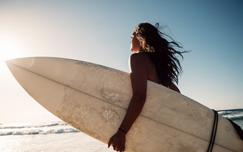 Woman with long brown hair holding a white surfboard walking along the beach at sunset.
