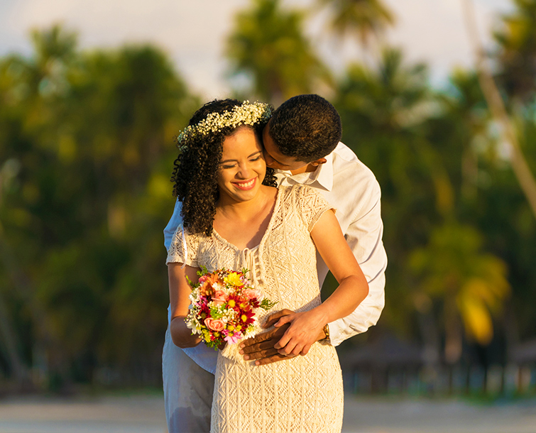 A smiling bride being hugged by husband while holding a bouquet of colorful flowers.