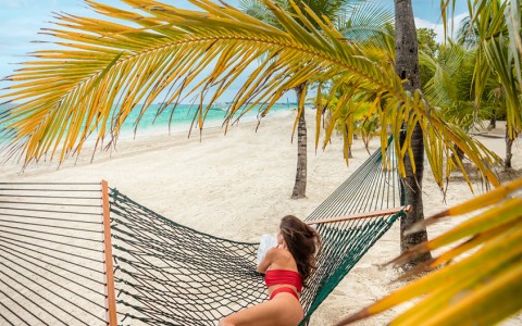 Woman wearing a red bikini laying down in a green hammock on the beach looking at the bright blue ocean.