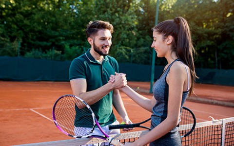 Male and female shaking hands on the dark orange tennis court with the sun shining from above.