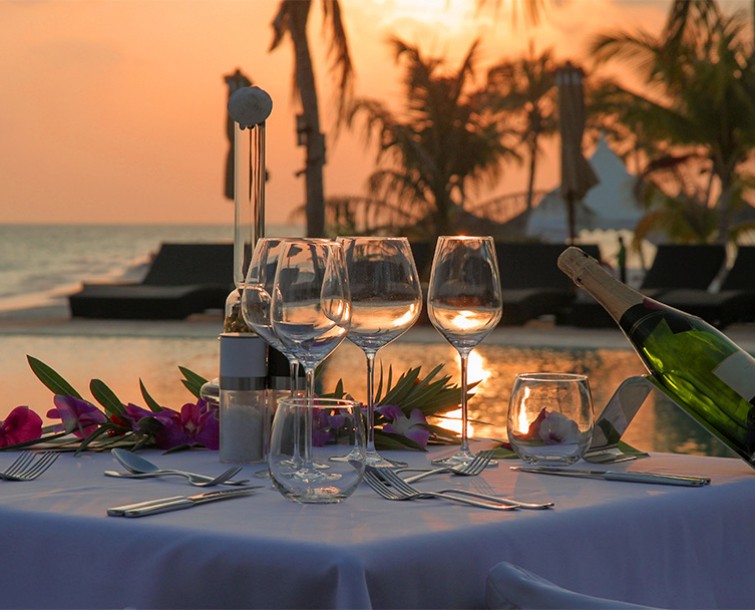 A white linen table set up for dinner service next to the water at sunset with palm trees in the distance.