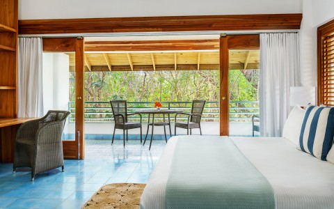Side view of inside the room with a light blue and white covered bed and dining table located on the balcony with big green trees in the distance.