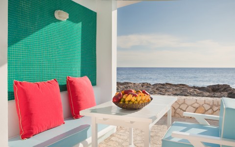 White outdoor table with chairs and two red throw pillows located next to the blue ocean.