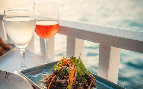 Seared meat garnished with shredded carrot and parsley on light blue plate and a glass of rose on the table next to the ocean at sunset.