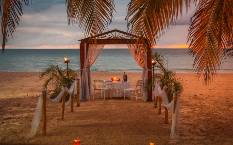 A table on the beach at sunset setup for dinner surrounded by candles and palm trees. 