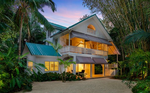 Two story home dimly lit surrounded by tall palm trees and green plants.