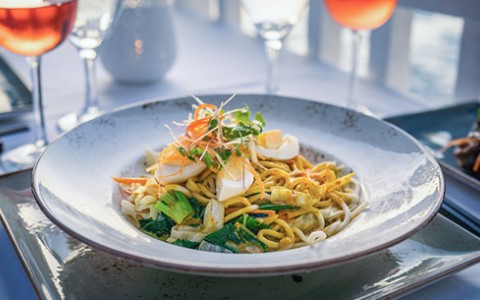 A large bowl of sauteed noodles topped with eggs and scallions served with a glass of rose.