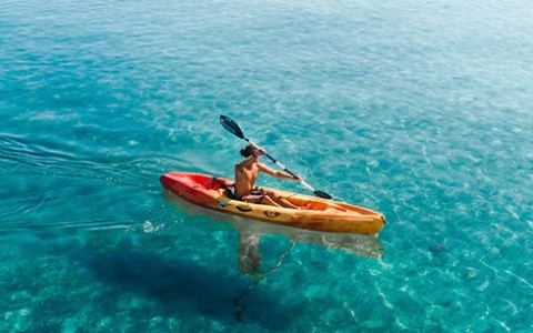 A male kayaking in the blue water with the sun shining.