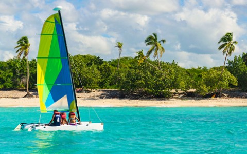 Neon blue, green, and yellow sailboat with three people on it in the blue water.